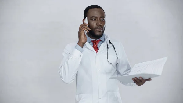 African american doctor in white coat talking on cellphone while holding folder with documents — Stock Photo