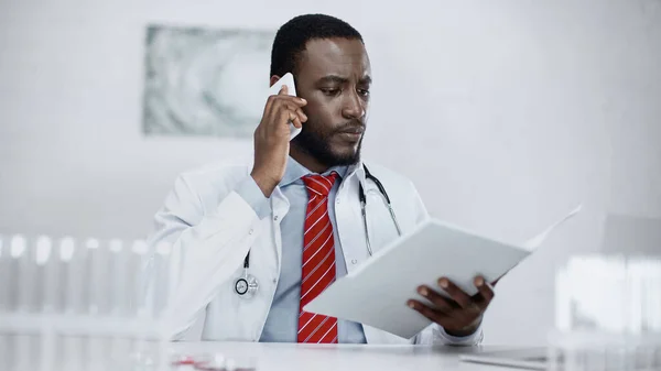 Serious african american doctor talking on smartphone and holding paper folder — Stock Photo