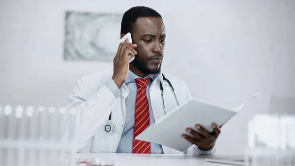 Médico afroamericano serio hablando en el teléfono inteligente y la celebración de la carpeta de papel - foto de stock