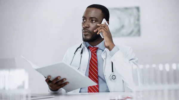 Seriöser afrikanisch-amerikanischer Arzt spricht mit Smartphone und hält Papiermappe in der Klinik — Stockfoto