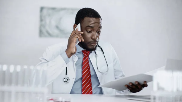 Serious african american doctor talking on cellphone and holding paper folder — Stock Photo
