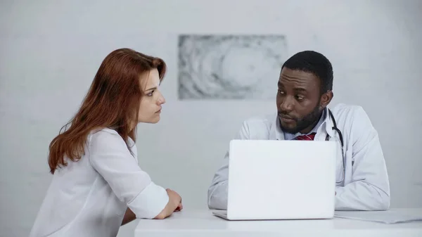 Afrikanisch-amerikanischer Arzt spricht mit rothaarigem Patienten in der Nähe von Laptop auf dem Schreibtisch — Stockfoto