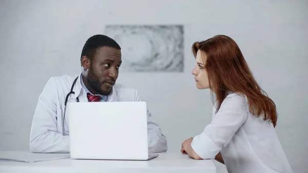 Bärtiger afrikanisch-amerikanischer Arzt im Gespräch mit rothaarigem Patienten in der Nähe von Laptop auf dem Schreibtisch — Stockfoto