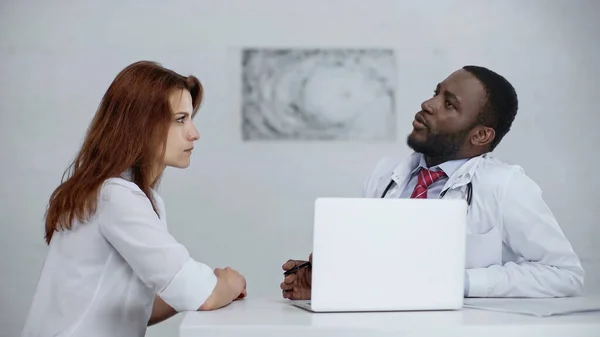 Africano americano médico hablando con pelirroja mujer cerca de portátil en escritorio - foto de stock