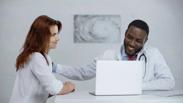 Alegre Africano americano médico falando com ruiva paciente perto laptop na mesa — Fotografia de Stock