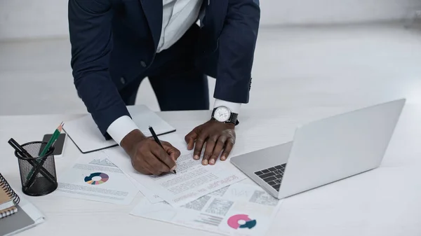 Частичный взгляд африканского американского бизнесмена, подписывающего документ рядом с ноутбуком на столе — стоковое фото