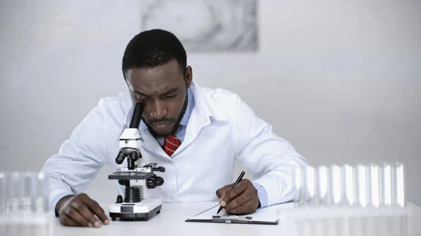 African american scientist looking through microscope and writing on clipboard — Stock Photo