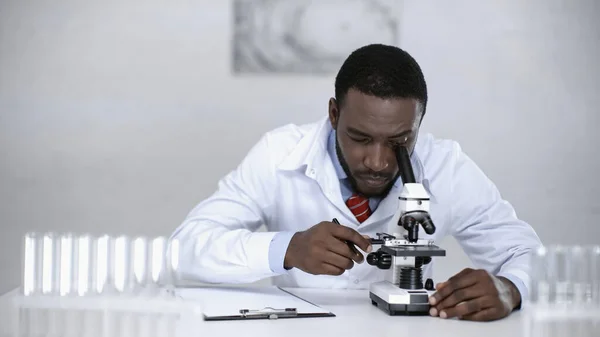 African american scientist looking through microscope near clipboard in lab — Stock Photo