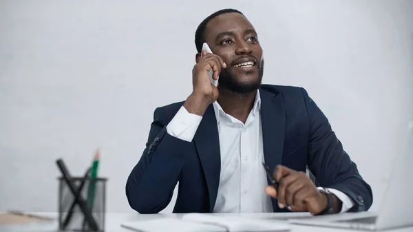 Hombre de negocios afroamericano feliz hablando en el teléfono inteligente cerca de la computadora portátil en el escritorio aislado en gris - foto de stock