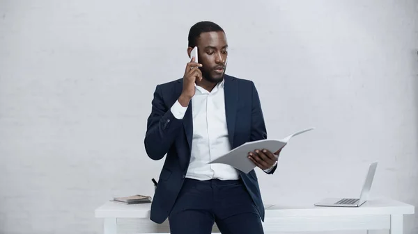 Hombre de negocios afroamericano serio hablando en el teléfono inteligente mientras está de pie con la carpeta - foto de stock