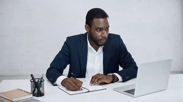 Serious african american businessman writing on notebook while looking at laptop on grey — Stock Photo