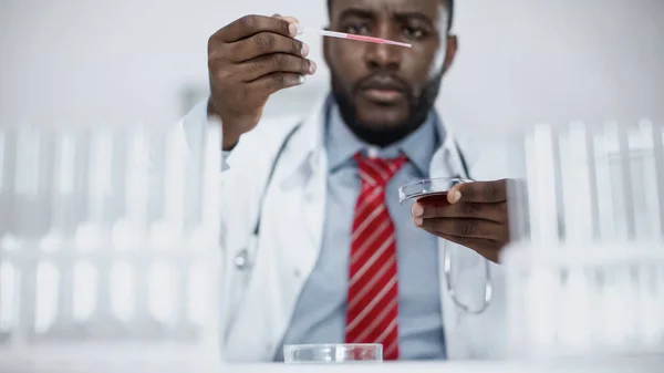 Científico afroamericano borroso mirando la pipeta con líquido rojo en el laboratorio - foto de stock