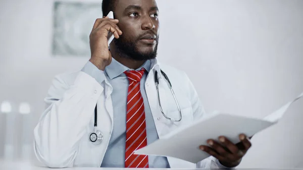 African american doctor talking on mobile phone while holding blurred folder — Stock Photo