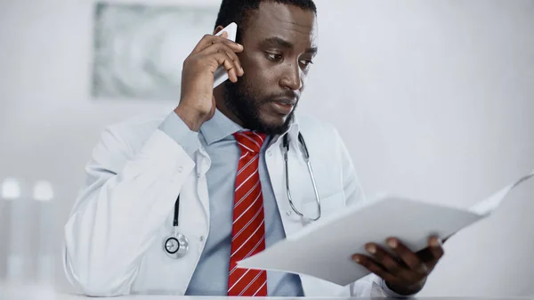African american doctor talking on smartphone while holding blurred folder — Stock Photo