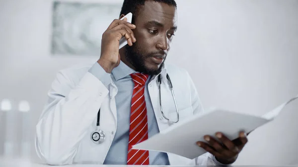 Afrikanisch-amerikanischer Arzt spricht auf Smartphone, während er verschwommenen Ordner hält — Stockfoto