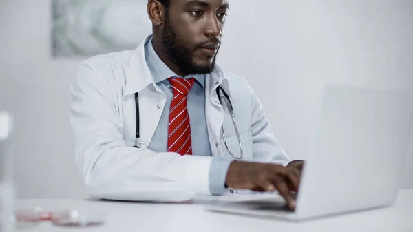 Barbudo afroamericano médico escribiendo en el ordenador portátil en la clínica - foto de stock