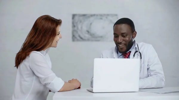 Glücklich afrikanisch-amerikanische Arzt im Gespräch mit rothaarigen Patienten in der Nähe von Laptop auf dem Tisch — Stockfoto