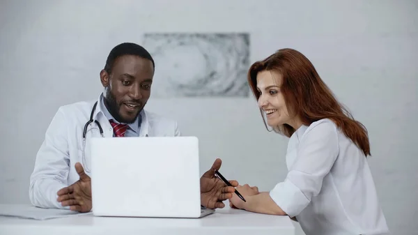 Africano americano medico parlando con allegro rossa paziente vicino laptop su tavolo — Foto stock