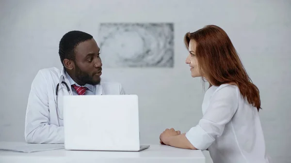 Afro americano medico parlando con compiaciuto rossa paziente vicino laptop su tavolo — Foto stock