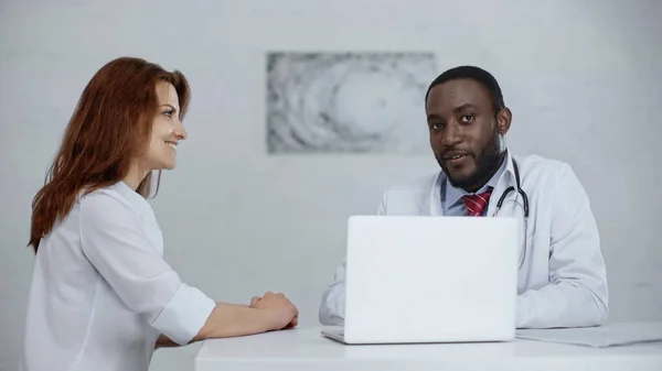 Médico afroamericano hablando con un paciente positivo cerca de la computadora portátil en la mesa - foto de stock