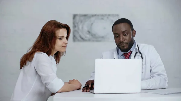 Africano americano médico y pelirroja paciente mirando portátil en mesa - foto de stock