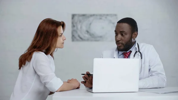 Africano americano médico hablando con pelirroja mujer cerca de portátil en mesa - foto de stock