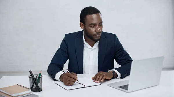 Hombre de negocios afroamericano serio mirando a la computadora portátil mientras sostiene la pluma cerca del cuaderno en gris - foto de stock