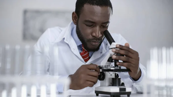 African american scientist in white coat looking through microscope in laboratory — Stock Photo