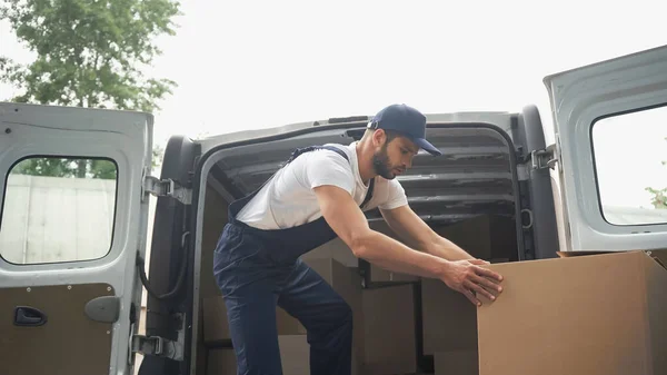 Vista lateral del mensajero en caja de cartón de sujeción uniforme cerca del automóvil al aire libre - foto de stock