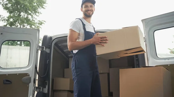 Lächelnder Kurier mit Karton und Blick auf Kamera in der Nähe von Auto im Freien — Stockfoto