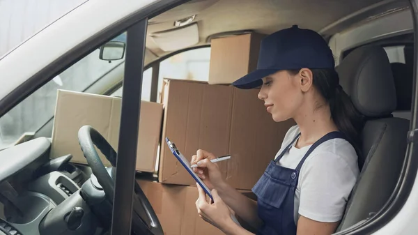 Vue latérale de la livraison femme écriture sur presse-papiers près des boîtes dans la voiture — Photo de stock