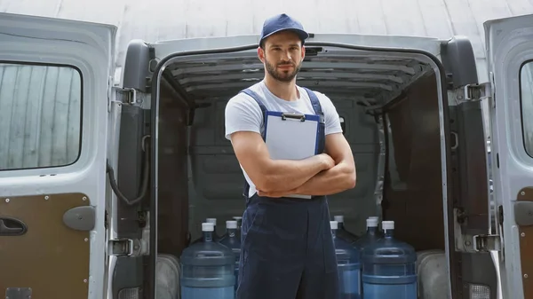 Mensajero en uniforme sujetando el portapapeles y mirando a la cámara cerca de botellas en el coche al aire libre - foto de stock