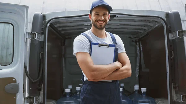 Smiling courier with clipboard looking at camera near bottles in car outdoors — Stock Photo