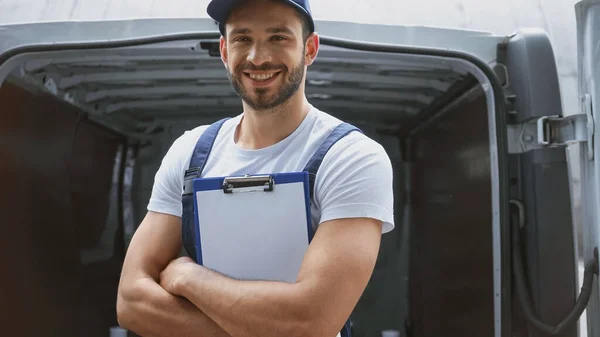 Cheerful delivery man crossing arms and holding clipboard near car outdoors — Stock Photo