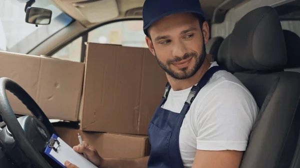 Lächelnder Kurier mit Klemmbrett sitzt in Auto neben Kartons — Stockfoto