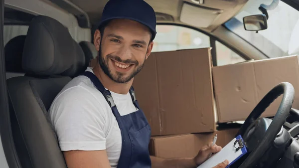 Positiver Kurier hält Klemmbrett und blickt auf Kamera in der Nähe von Boxen im Auto — Stockfoto