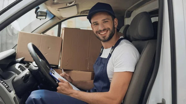 Lächelnder Kurier in Uniform hält Klemmbrett in der Nähe von Boxen im Auto — Stockfoto