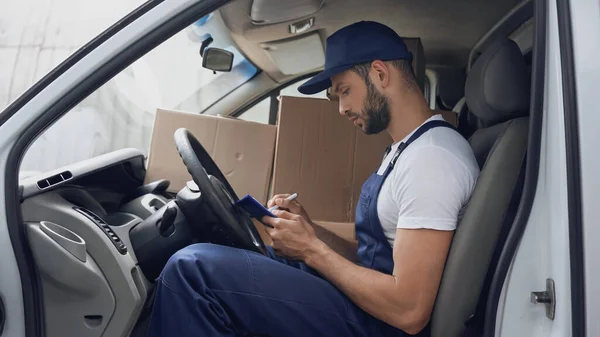 Side view of delivery man writing on clipboard near carton boxes in car — Stock Photo