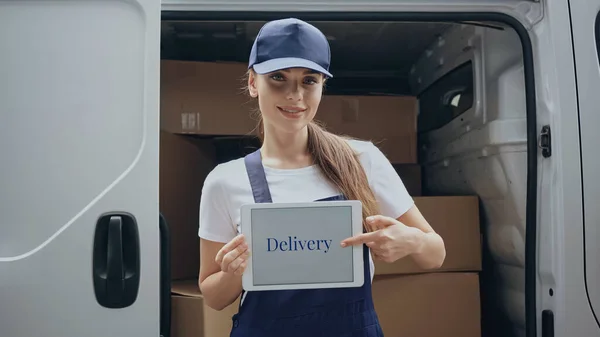 Courrier souriant pointant vers la tablette numérique avec lettrage de livraison près de la voiture à l'extérieur — Photo de stock