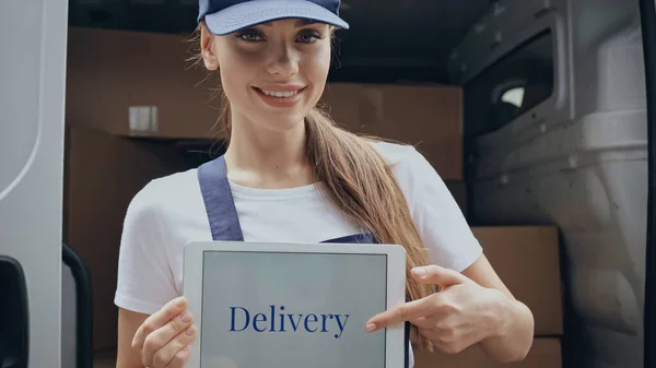 Positive delivery woman pointing at digital tablet with lettering near blurred car outdoors — Stock Photo