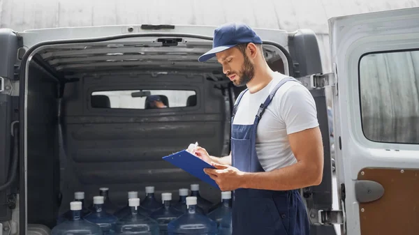 Courier in cap writing on clipboard near car outdoors — Stock Photo