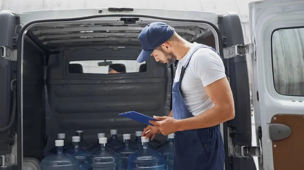 Seitenansicht des Lieferanten in Overalls mit Klemmbrett in der Nähe von Wasserflaschen im Auto — Stockfoto