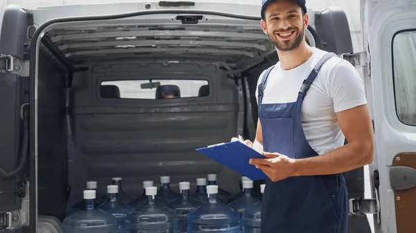 Mensajero alegre en overoles escribiendo en el portapapeles cerca de coche al aire libre - foto de stock