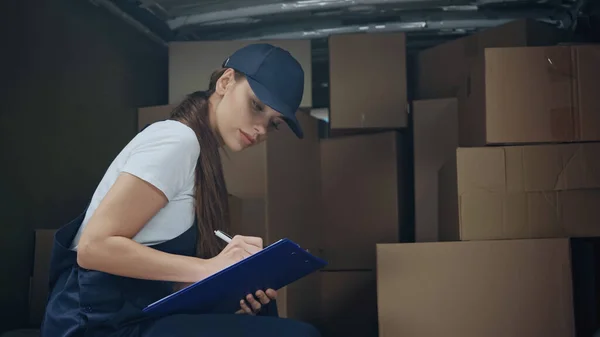 Young deliverywoman writing on clipboard near boxes in car outdoors — Stock Photo