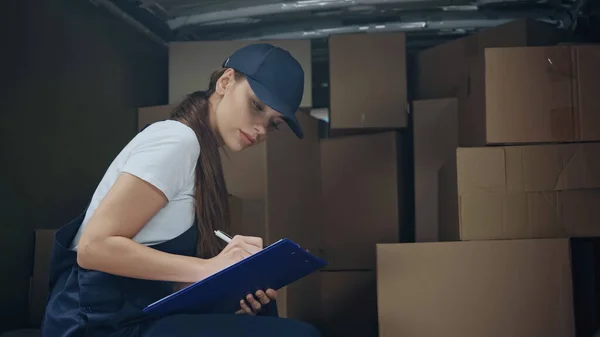 Jeune livreuse écrivant sur presse-papiers près des boîtes en voiture à l'extérieur — Photo de stock