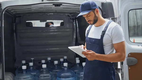 Mensajero en overoles usando tableta digital cerca de botellas en auto al aire libre - foto de stock