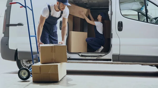 Courier sostiene la caja de cartón cerca del carro y colega en el coche al aire libre - foto de stock