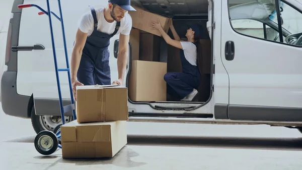 Courier holding carton box near cart and colleague in car outdoors — Stock Photo