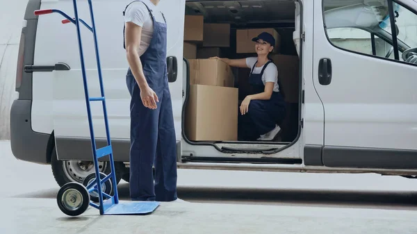 Mujer de entrega sonriente que trabaja con cajas de cartón y colega cerca del automóvil al aire libre - foto de stock