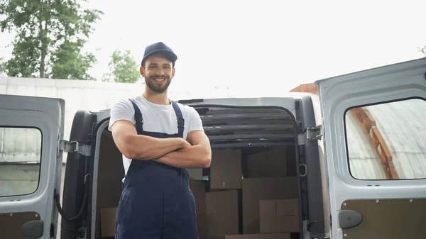 Smiling courier in cap and overalls looking at camera near boxes in car outdoors — Stock Photo